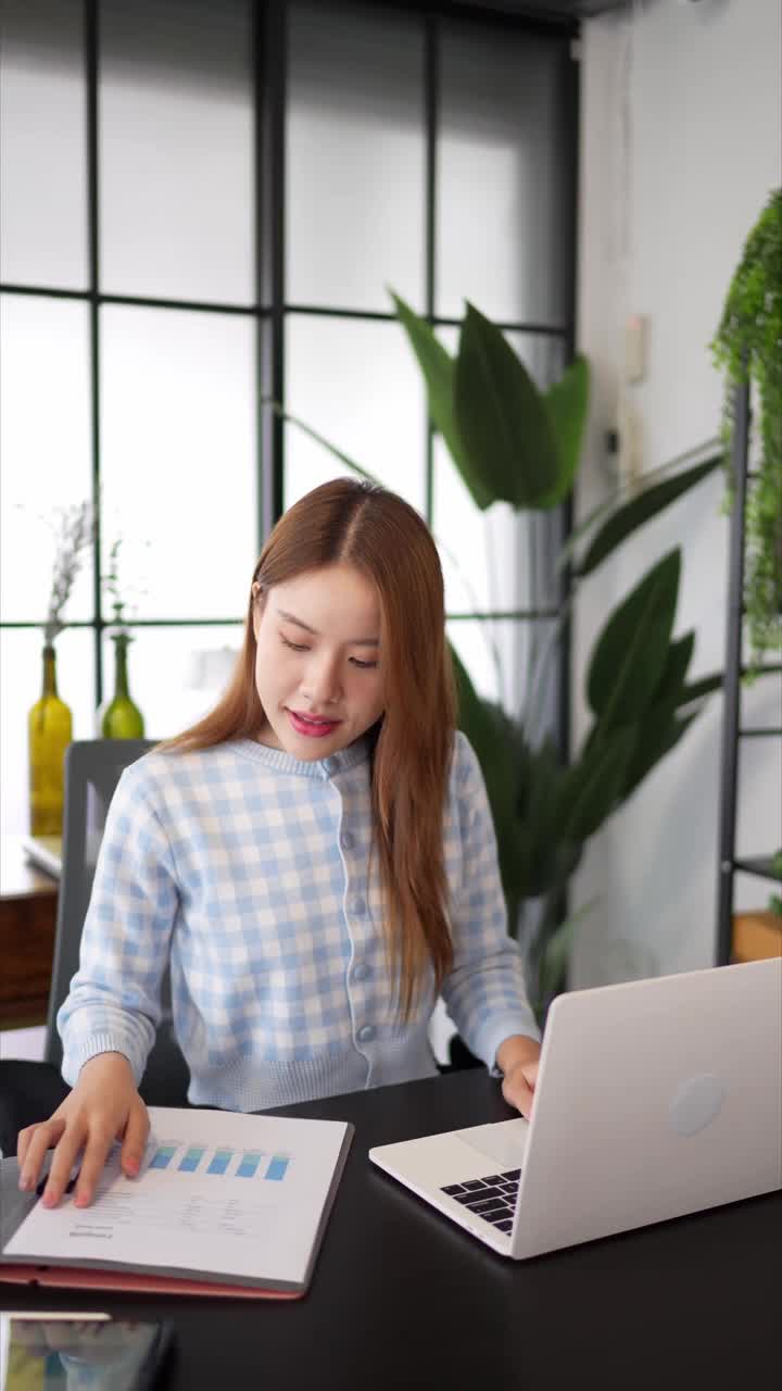 一位穿着格子衬衫、自信的女商人在一间以大窗户和绿色植物为背景的时髦办公室里，一边看文件，一边用笔记本电脑工作视频素材