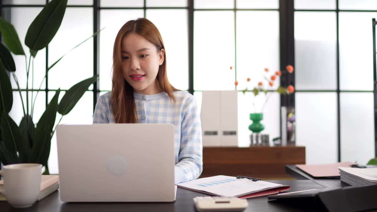 一位穿着格子衬衫、自信的女商人在一间以大窗户和绿色植物为背景的时髦办公室里，一边看文件，一边用笔记本电脑工作视频素材