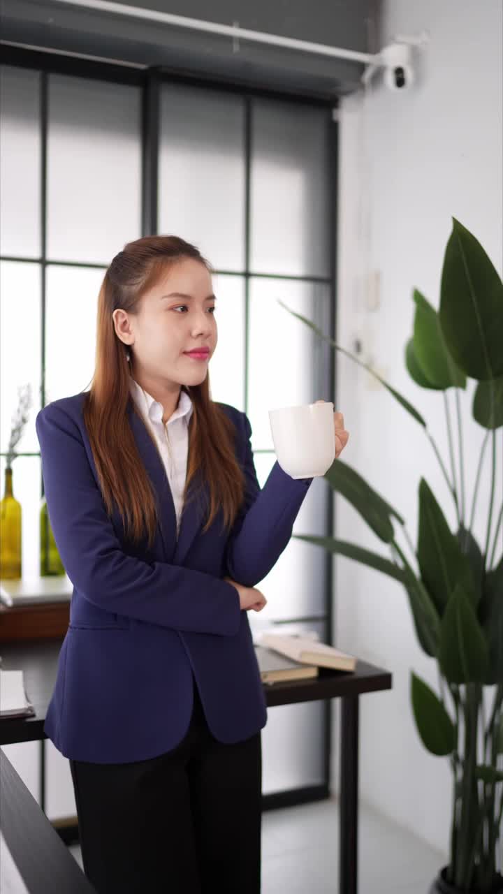 一位身穿海军蓝西装的女商人站在一间现代化的办公室里，手里拿着一个白色的咖啡杯，看上去若有所思，周围是办公室里的植物和放着文件的架子视频素材