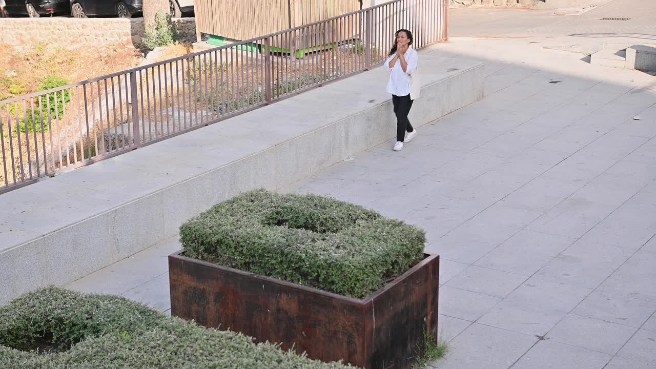 一名女子穿着白衬衫，肩上背着包，走在城市公园的户外打手机视频素材