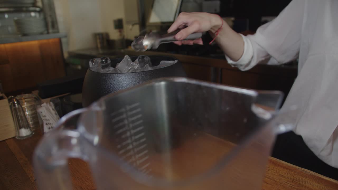 一个美丽的慢动作镜头，一个年轻的女清洁工用钳子夹起一桶冰，准备在一家餐厅的吧台上调制鸡尾酒。视频素材