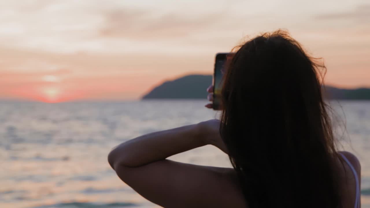 日落时分，一名女子在海滩上用智能手机自拍视频素材