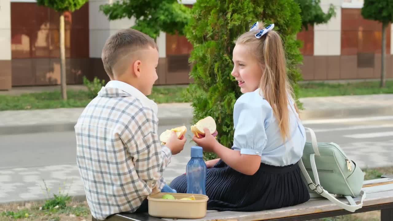 学校吃饭，孩子们女孩和男孩小学生在街上吃午饭用汉堡包，孩子们哥哥姐姐上学前吃三明治，快餐学生视频下载