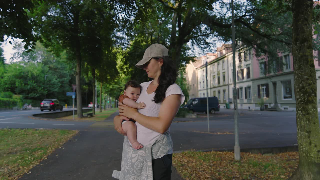 母亲抱着她的孩子站在绿树成荫的安静街道上，戴着棒球帽，在绿树成荫的城市环境中，这是一个和平的时刻视频素材