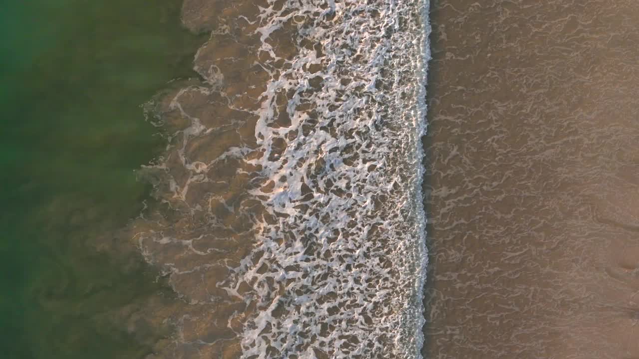 柔和的海浪冲刷到沙滩上，清澈的绿色海水与棕色海岸线相遇，形成美丽的自然图案，鸟瞰图视频素材