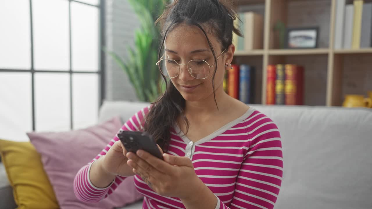 一位身穿条纹衬衫、戴着眼镜的年轻西班牙裔女子在室内盯着智能手机。视频素材