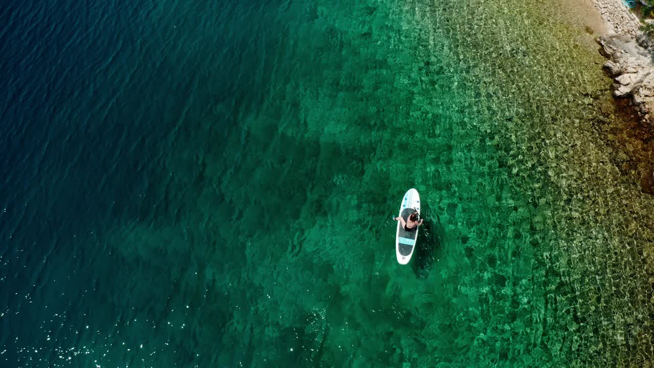 俯视图，一个女人在蔚蓝的大海中移动，坐在桨板上视频下载