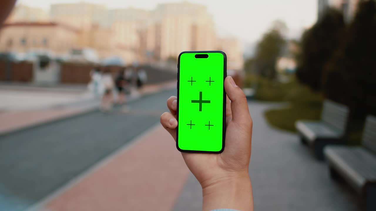 一只手拿着智能手机的特写镜头，以一个阳光明媚的日子里，城市街道的人行道为背景，模拟绿色屏幕。现代手机应用程序模型的概念视频下载