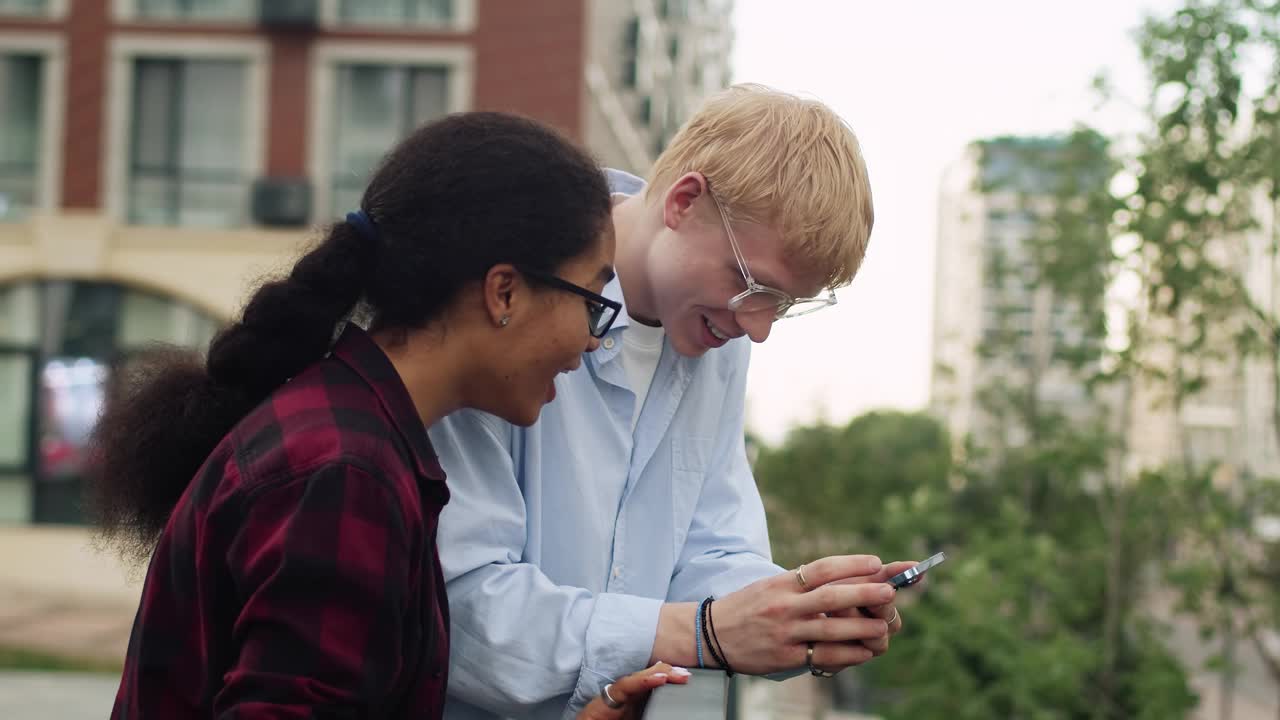 一个年轻的白人男孩和一个年轻的黑人女孩看着智能手机，变得非常高兴，在户外以建筑为背景大笑视频下载