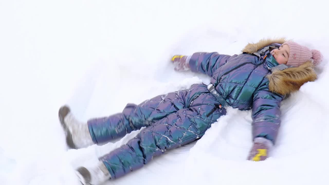 一个穿着蓝色连体衣的女孩在雪地里制作雪天使。冬季乐趣，户外游戏，圣诞节视频下载