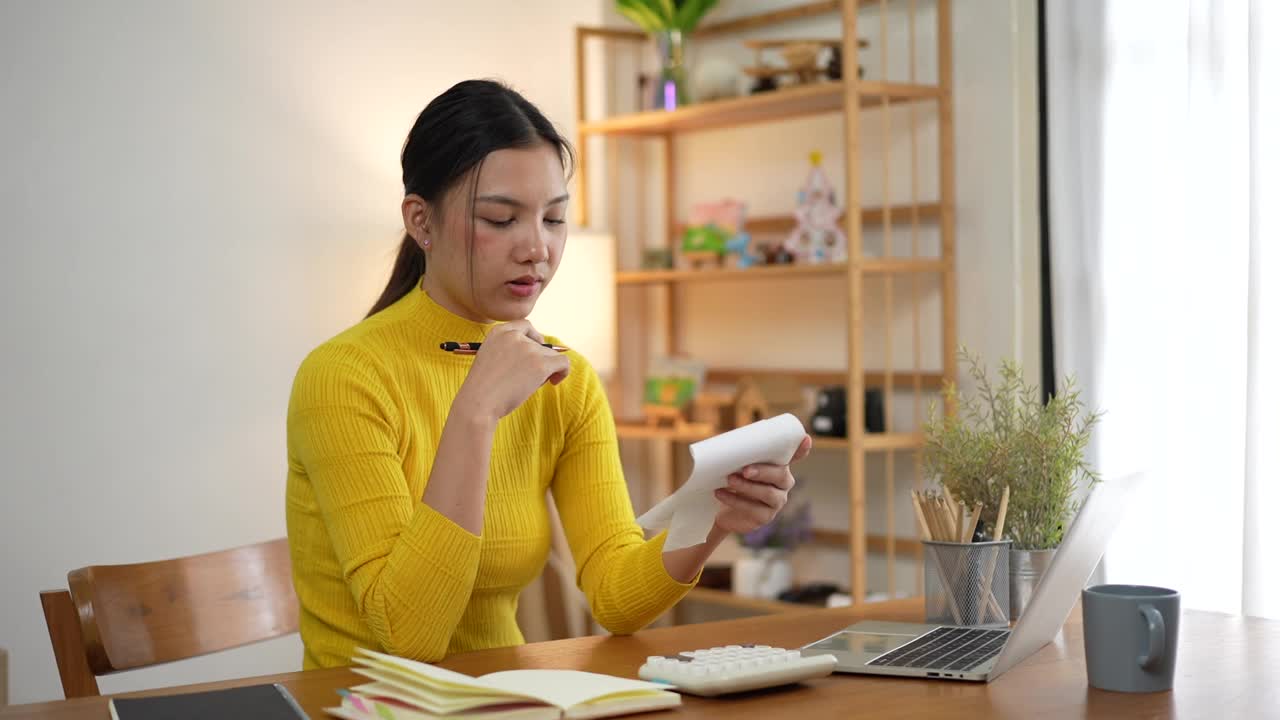 忧心忡忡的亚洲妇女看着纸质账单，检查保险发票或税务文件，计算费用，在家厨房用笔记本电脑工作视频下载