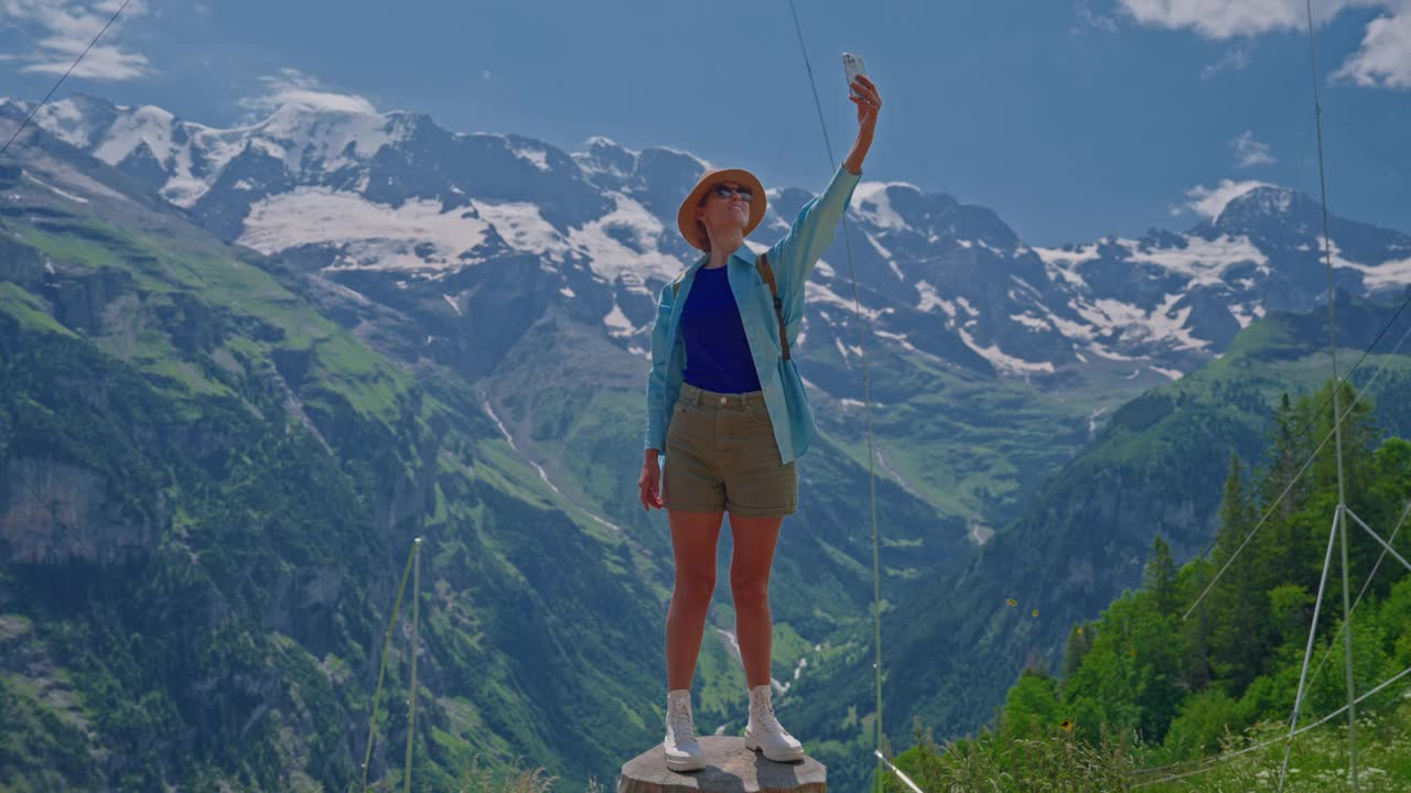 一个戴着帽子的女孩在瑞士山区用手机自拍，背景是美丽的阿尔卑斯山脉。视频下载