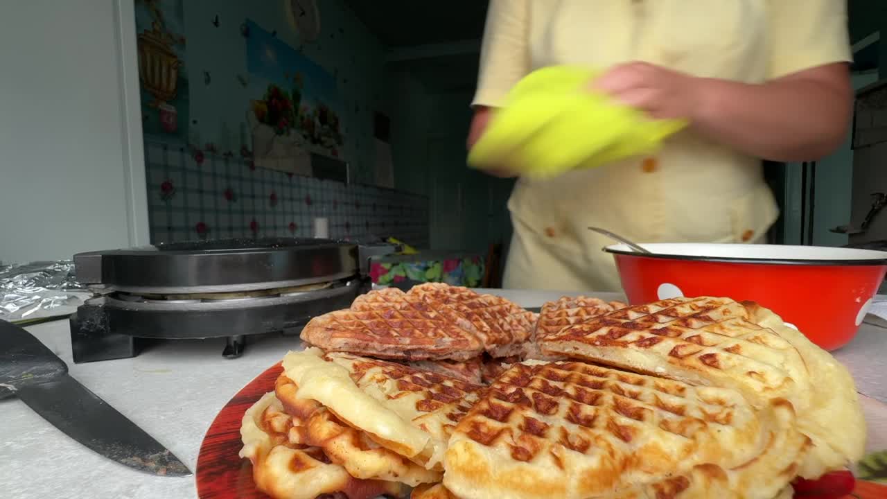 电子华夫饼制造机，发出蒸汽。旁边是准备好的华夫饼。一位妇女正在擦拭工作台面。视频素材