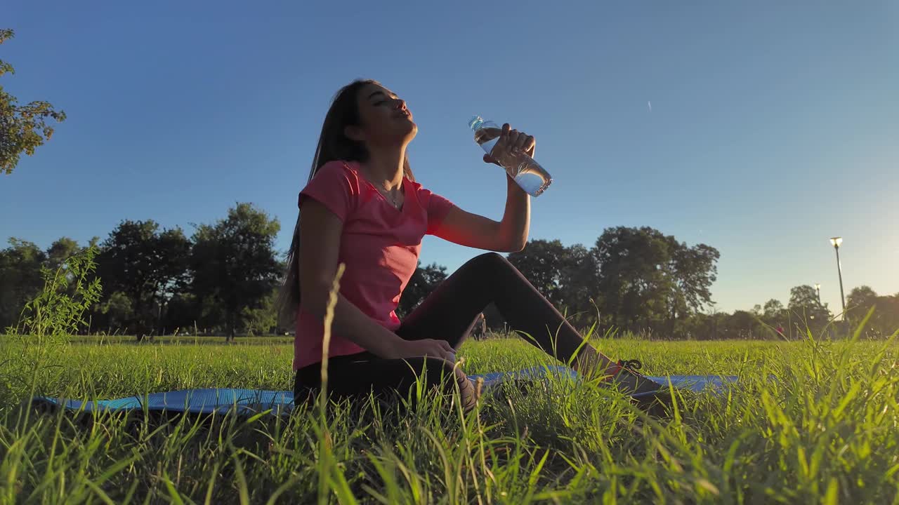 夏日日落时分，一位口渴的年轻女子在城市公园草地上的瑜伽垫上喝水和锻炼。视频下载