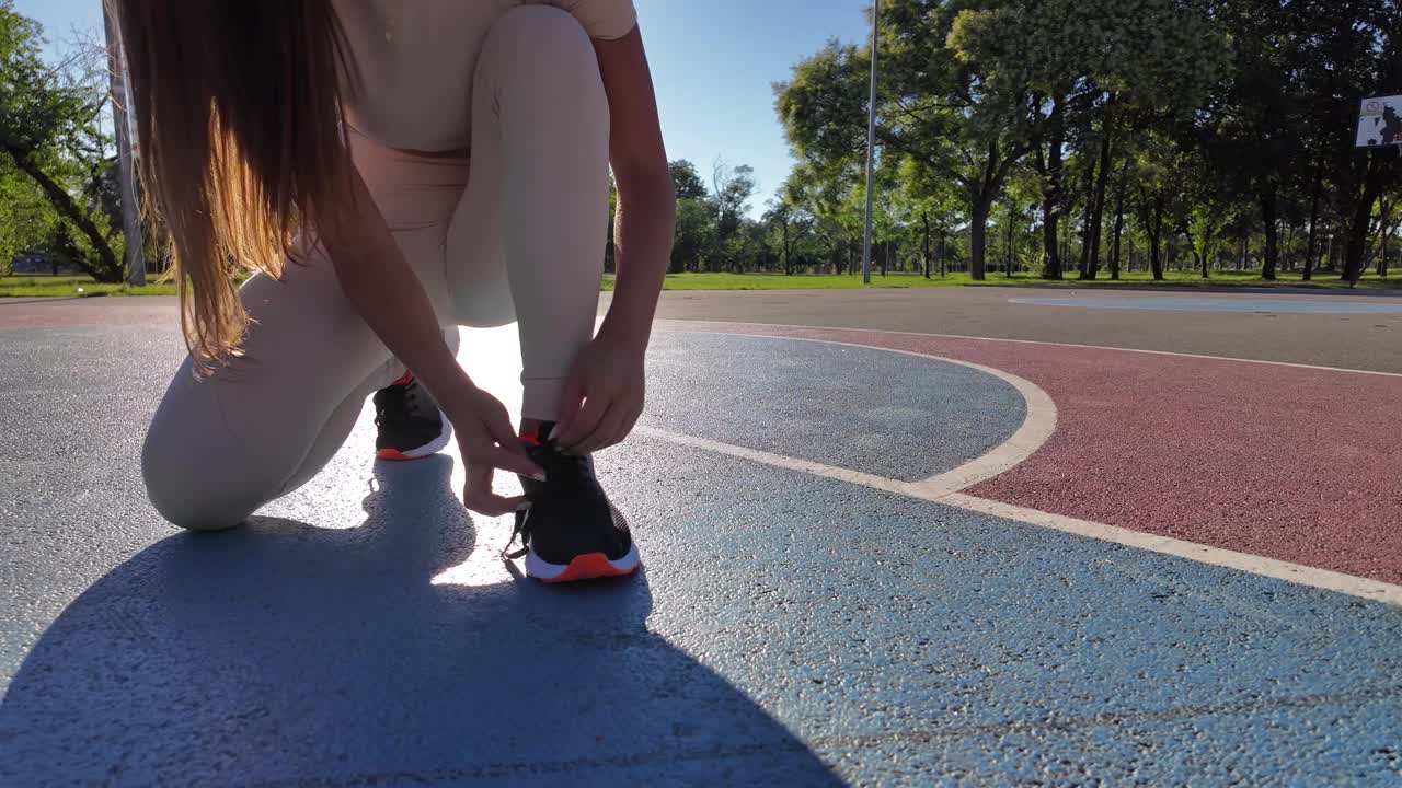 在夏日阳光明媚的日子里，运动的年轻女子在城市公园里系着跑鞋。视频下载