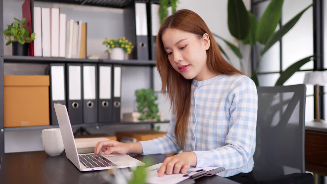 一位穿着格子衬衫、自信的女商人在一间以大窗户和绿色植物为背景的时髦办公室里，一边看文件，一边用笔记本电脑工作视频素材