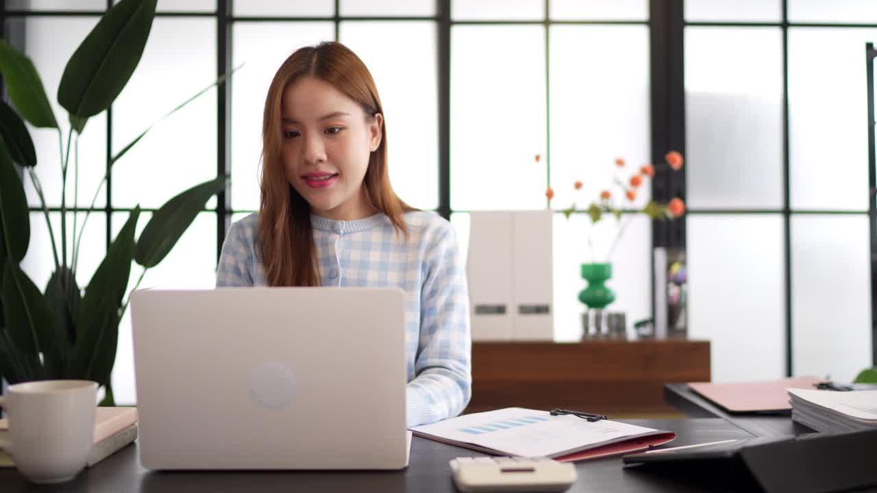 一位穿着格子衬衫、自信的女商人在一间以大窗户和绿色植物为背景的时髦办公室里，一边看文件，一边用笔记本电脑工作视频下载