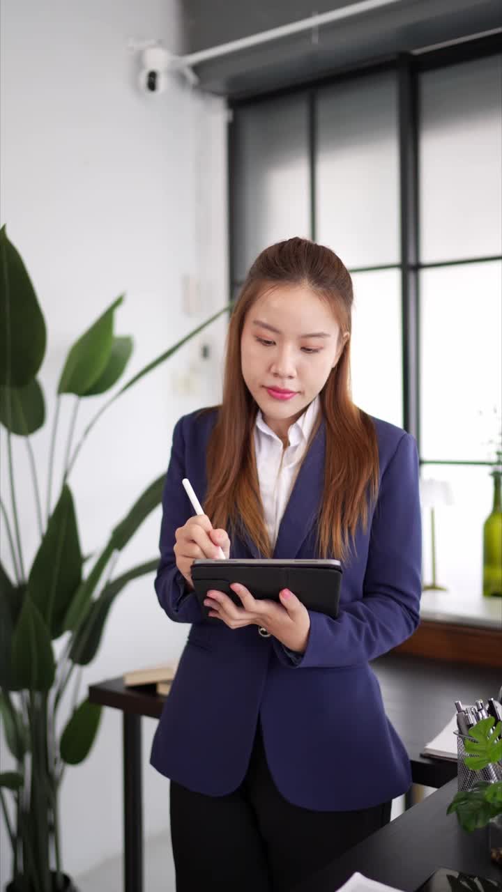 一位穿着海军蓝西装的自信女商人站在一间现代化的办公室里，用平板电脑做笔记，神情专注，周围是办公室的装饰和文件视频素材