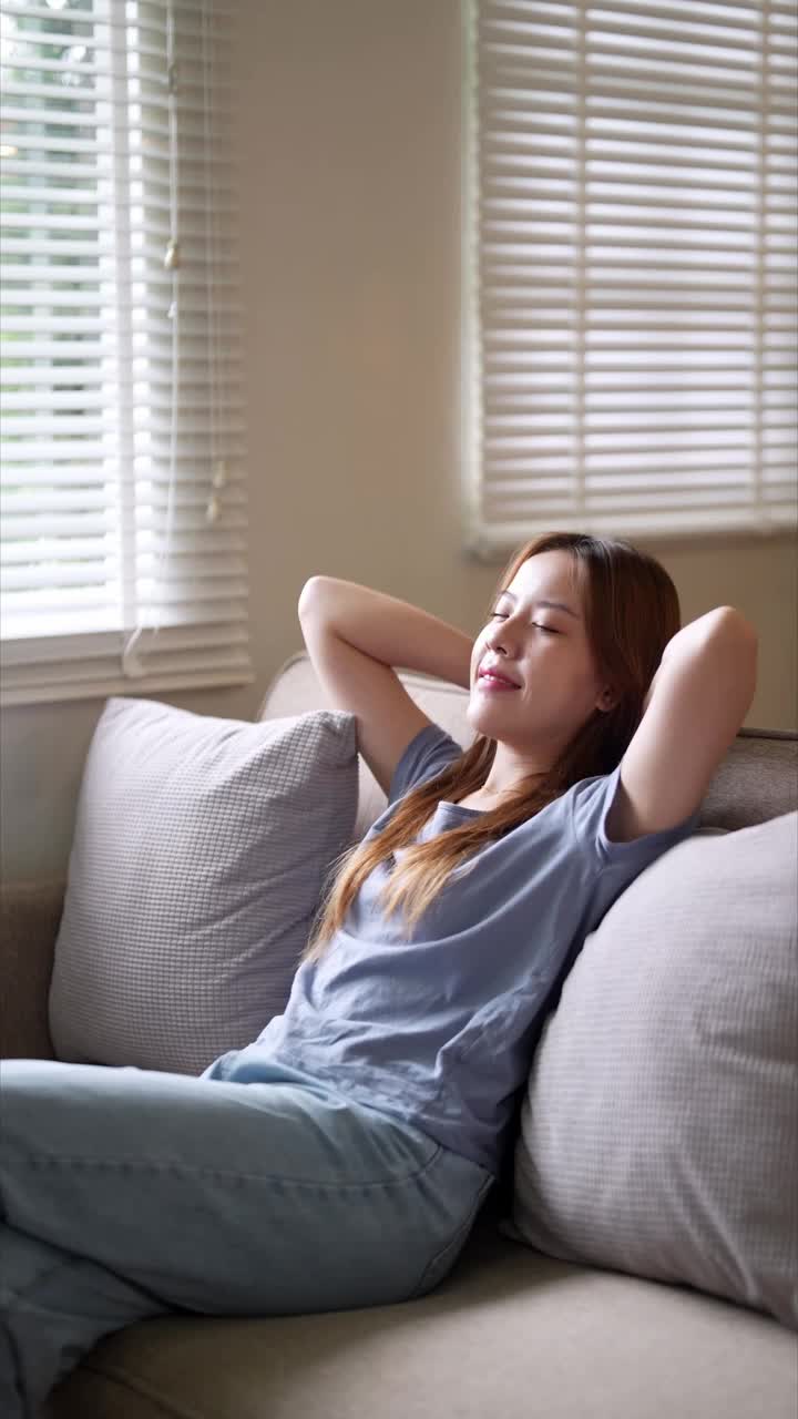 一名年轻女子躺在沙发上，双手放在脑后，闭着眼睛，脸上带着满足的微笑，享受着家里的宁静时光视频素材