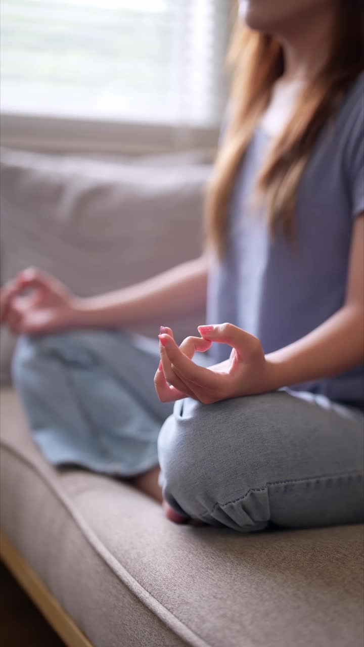 一个年轻的女人正盘腿坐在沙发上，闭着眼睛冥想，在一个平静而宁静的家庭环境中视频素材