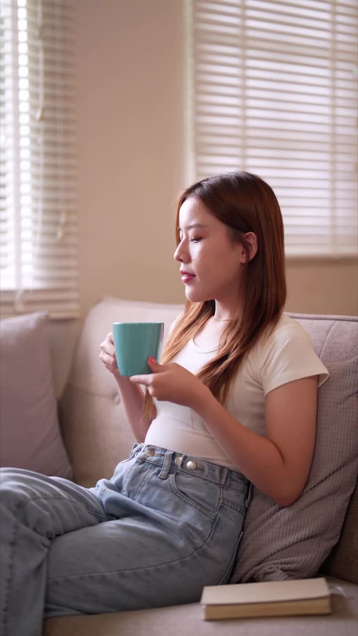 一个年轻女子坐在沙发上，端着杯子，若有所思地凝视着窗外宁静的室内环境视频素材