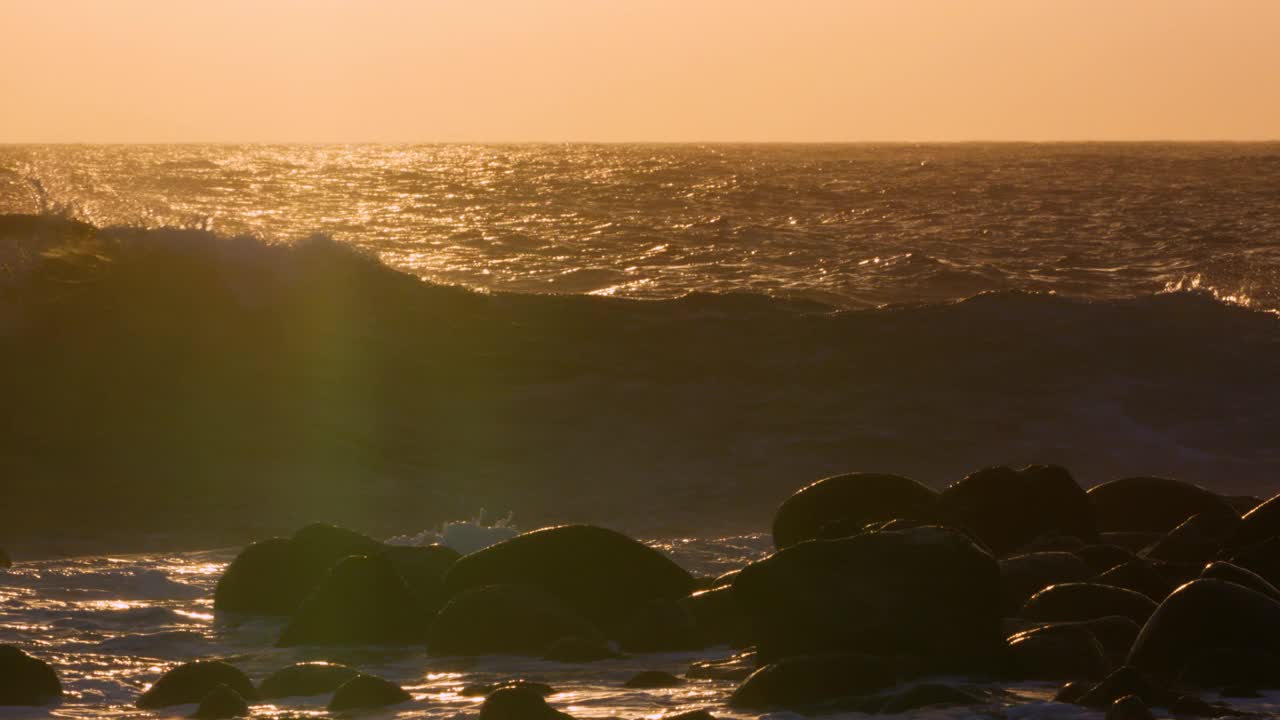 海浪拍打着黑色的海滩岩石，金色的夕阳在海面上闪闪发光。视频素材