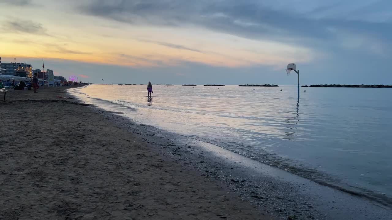 宁静的日落海滩场景，一个单独的篮球圈部分淹没在水里。人们沿着遥远的海岸线散步，温柔的海浪拍打着海岸视频素材