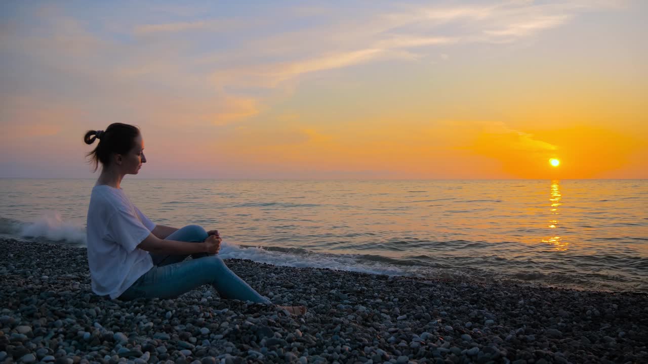 女人的肖像坐在海边，看着温暖的夕阳天空视频下载