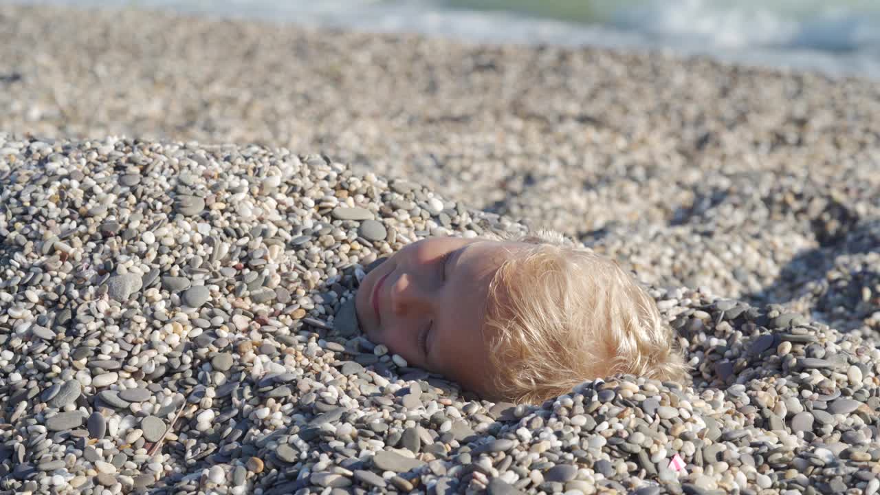 孩子们在沙滩上玩耍。快乐的孩子脸埋在沙子里的特写。视频下载