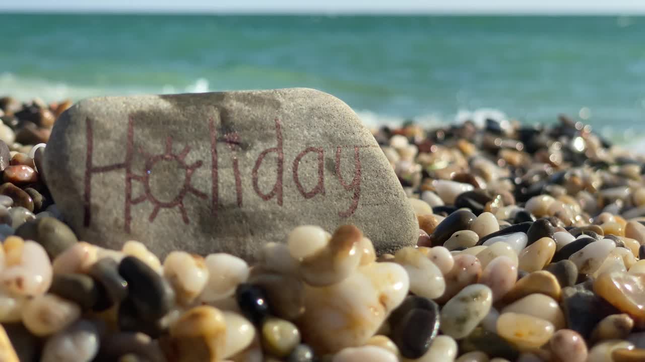 字假日手写在石头上，夏天的卵石沙滩被海浪溅起。夏季海滩度假背景。视频下载