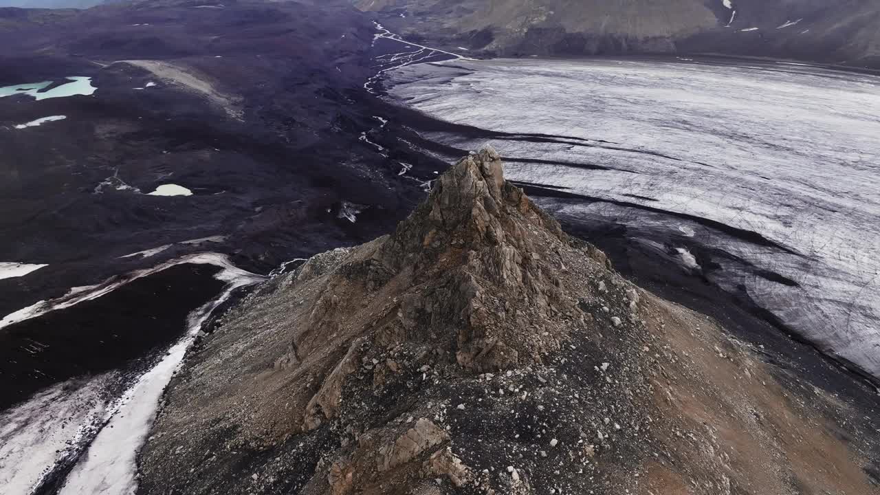 一座岩石山，周围是一片没有生命的田野，周围撒满了火山灰，旁边是一个巨大的冰川舌。冰岛景观鸟瞰图视频下载