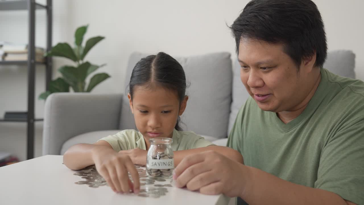 一个亚洲小女孩在家里和她的父亲一起学习投资积蓄，收集罐子里的硬币视频下载