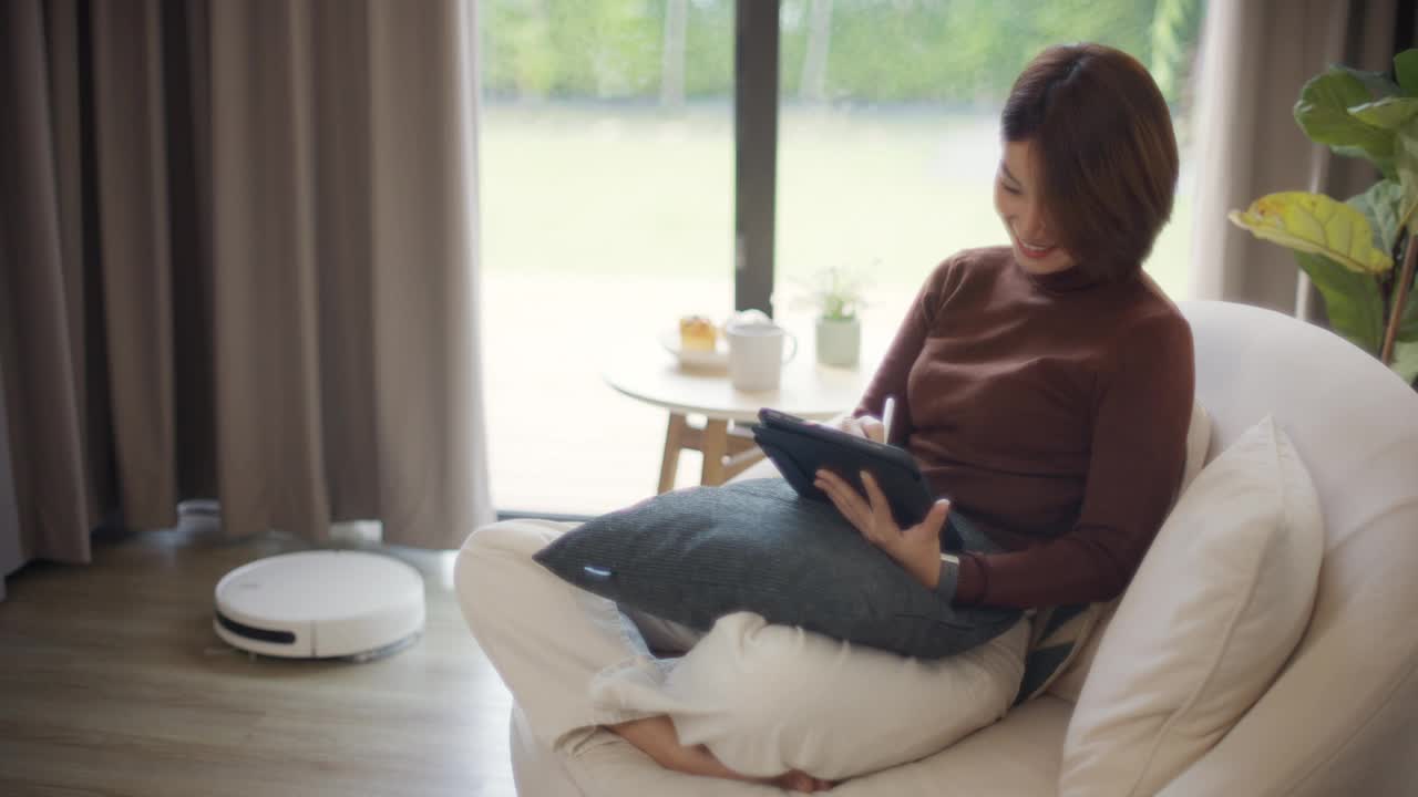 女人在家里摆弄数码平板电脑，机器人吸尘器在打扫地板视频素材