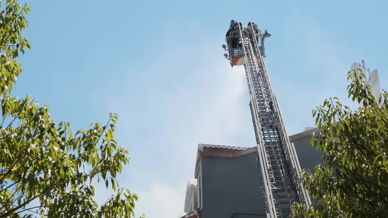 消防队员在高架平台上的后视图，用装有水管的加长梯扑灭燃烧的房屋，向带有烟雾的建筑物屋顶喷水。消防员灭火视频下载