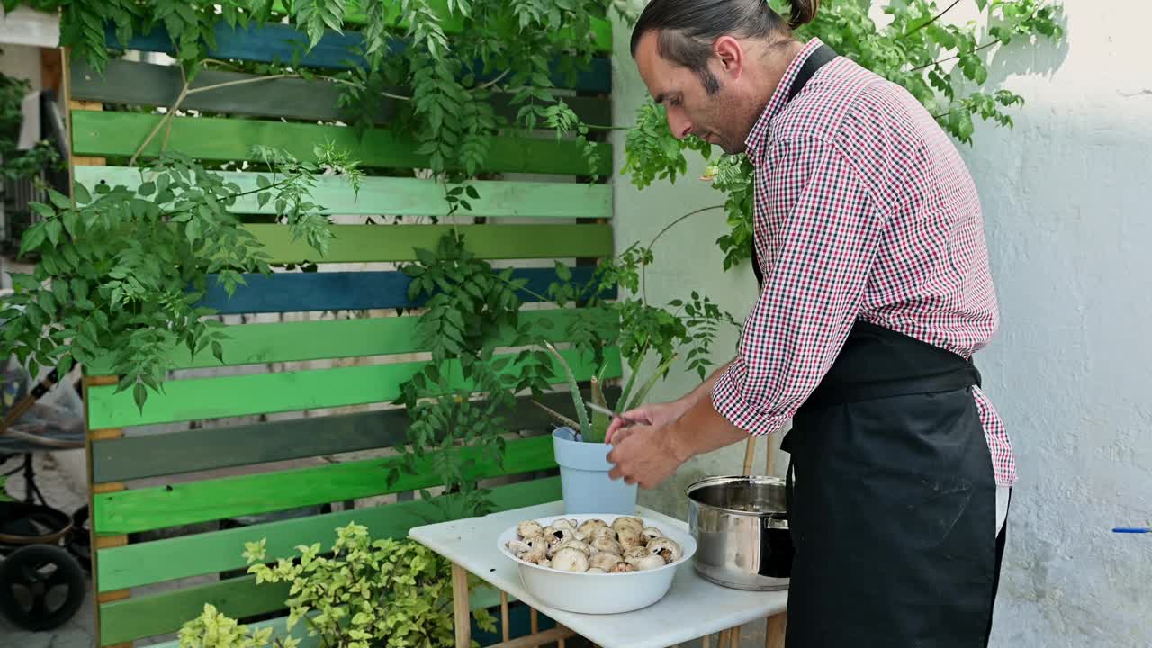 一名男子在郁郁葱葱的绿色植物和五颜六色的篱笆背景下的后院为烧烤准备蘑菇视频素材