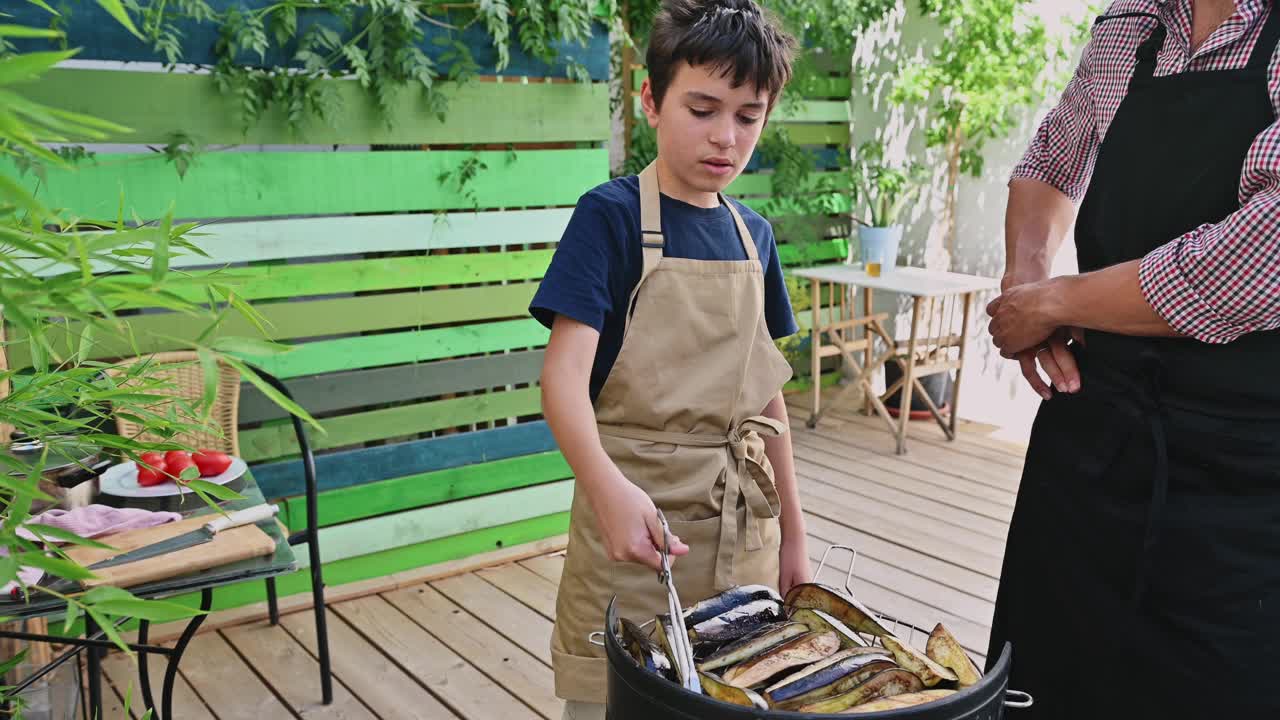 小男孩在阳光明媚的后院参加家庭烧烤，向穿着休闲围裙的成年人学习，创造了一种快乐的亲密体验。视频素材