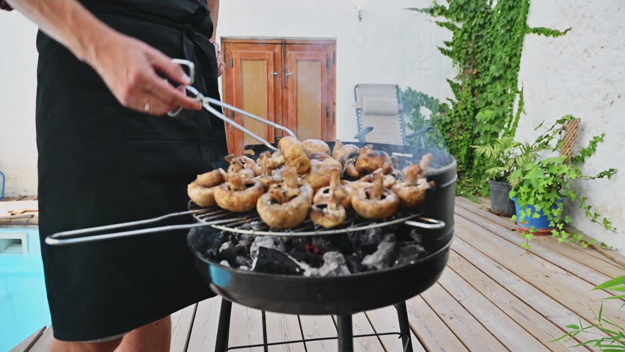 在一个休闲的户外聚会中，人们在后院的木炭烧烤架上烤蘑菇，周围是绿色植物和木制甲板。视频素材