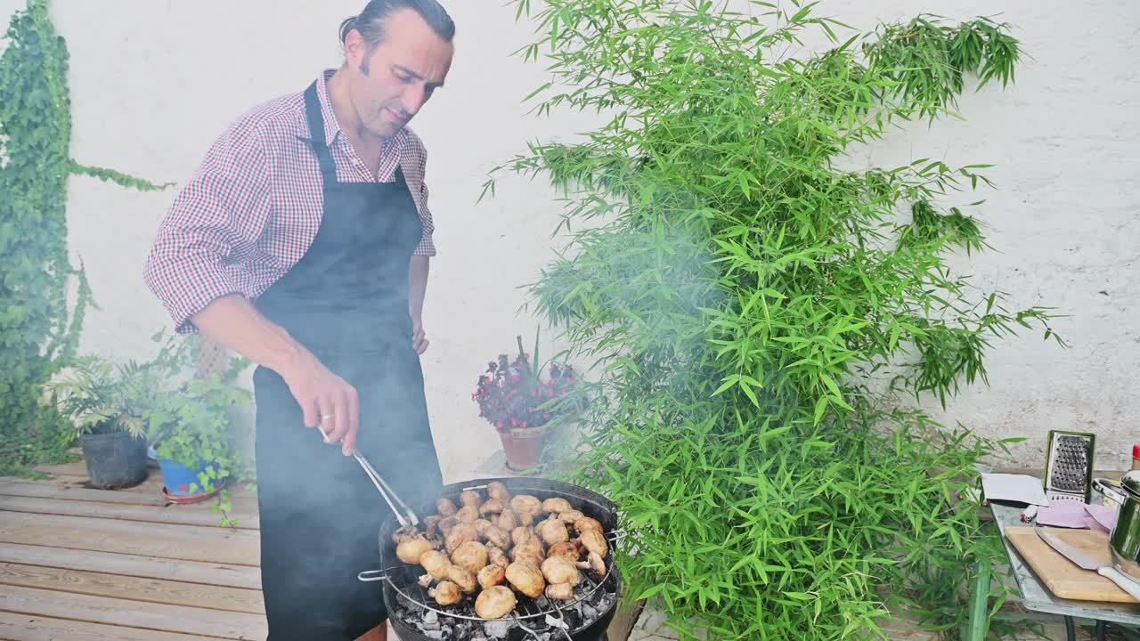 一名男子在长满生机勃勃的绿色植物的花园里烧烤土豆，穿着格子衬衫和围裙，享受着阳光明媚的户外烹饪体验视频素材