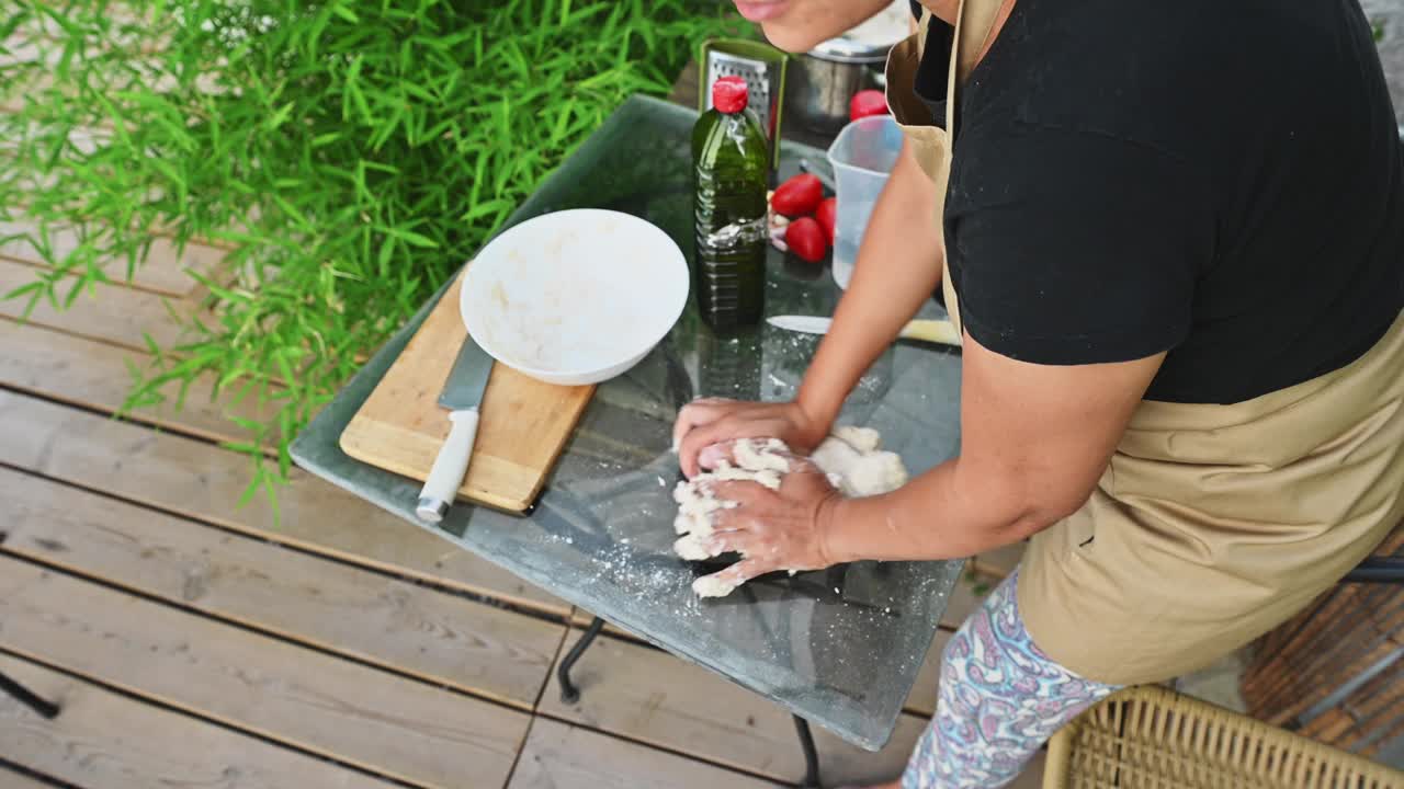 一名妇女在户外的玻璃桌上揉面，周围是花园中自制烹饪的食材视频素材