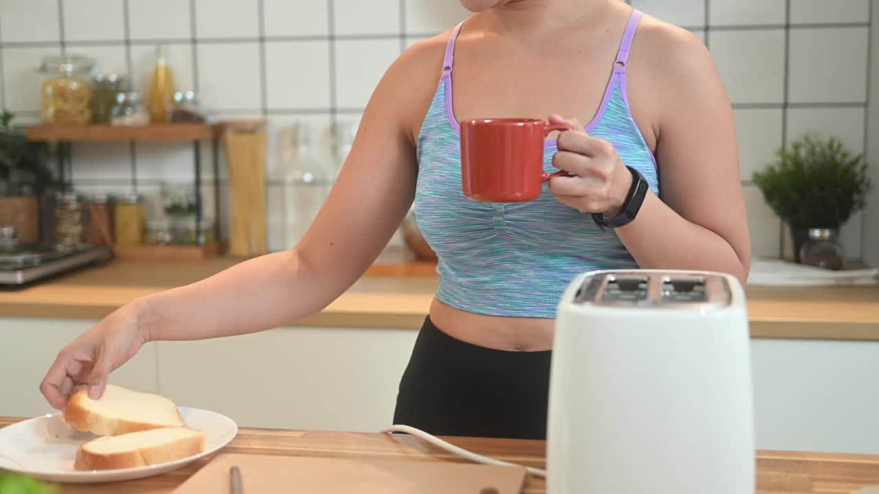 穿着运动服的年轻女子在厨房用烤面包机烤面包。视频素材