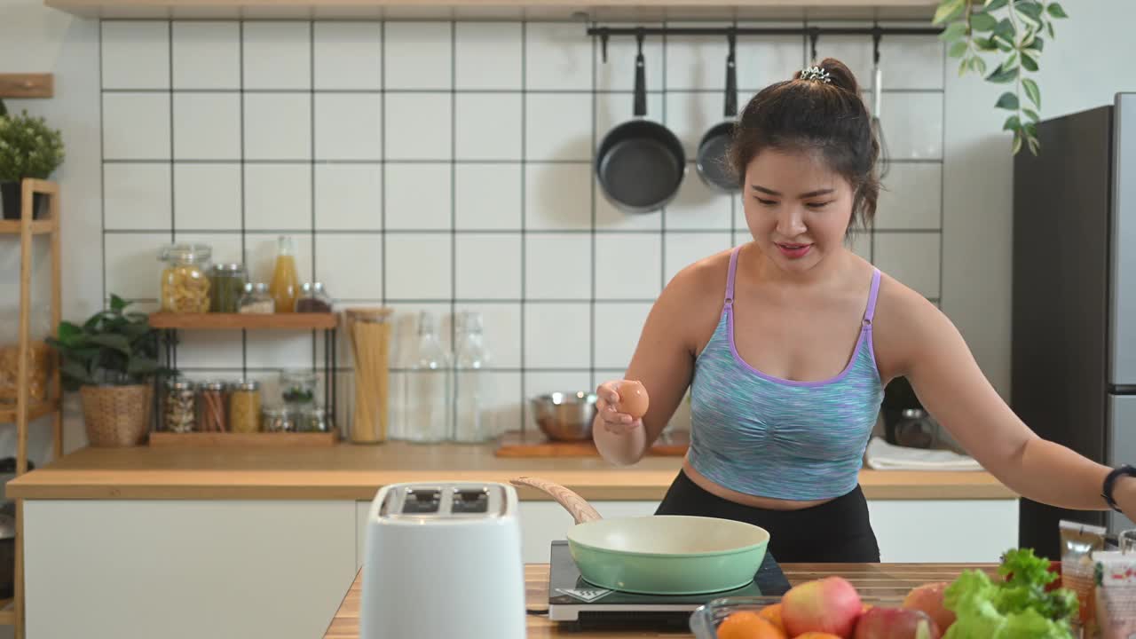 微笑的亚洲年轻女子穿着运动服在家庭厨房煎鸡蛋当早餐。视频素材