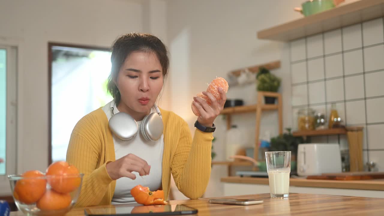 年轻漂亮的亚洲女人一边吃着新鲜的橘子一边在厨房柜台前摆弄着数码平板电脑。视频素材