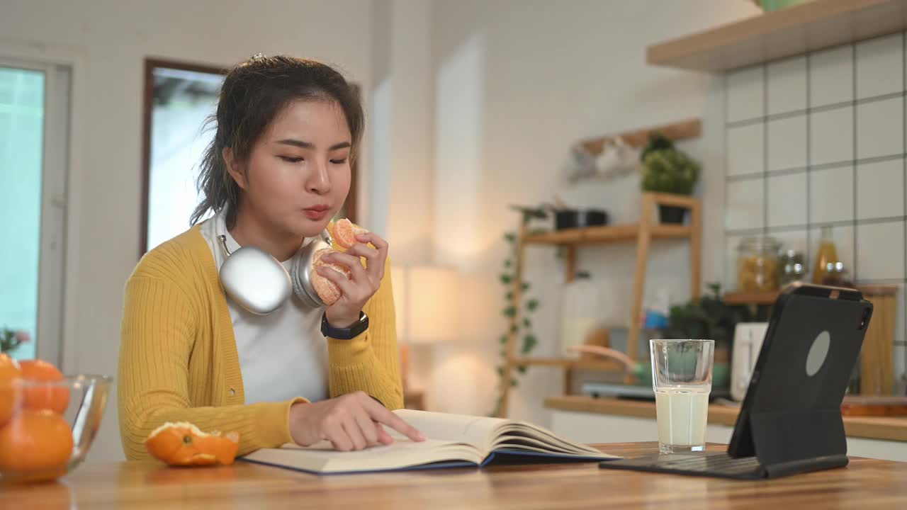 年轻的亚洲女人在厨房的柜台边看书边吃新鲜的橘子。视频下载