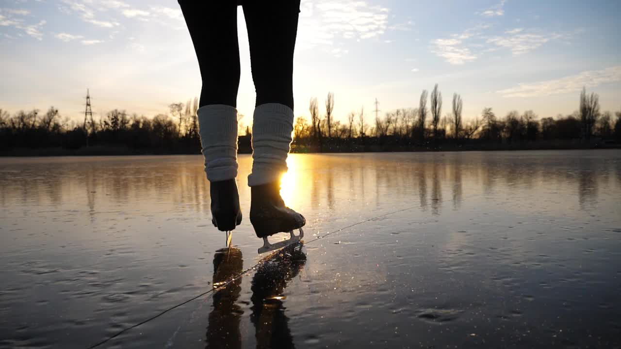 在美丽的夕阳下，滑冰者的女脚在结冰的河上滑冰。冰天雪地里，穿着花样滑冰鞋的女子在池塘上滑行。在冬天寒冷的天气里在湖边积极休闲的女运动员视频下载