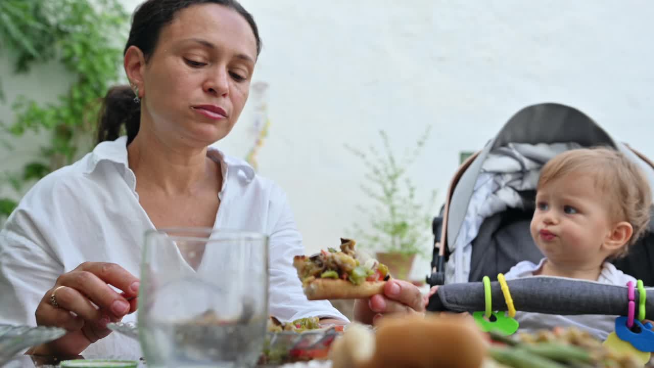 妈妈和宝宝在户外享受一顿饭，在宁静的花园环境中，用高脚椅和新鲜的食物捕捉片刻的亲密和家庭时光。视频素材