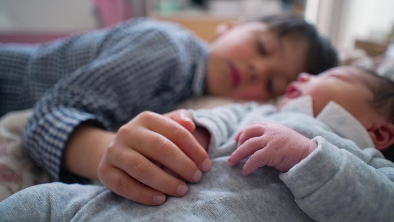 小男孩牵着刚出生的兄弟姐妹的手，躺在床上，微笑着，在一个充满爱和养育的家庭环境中，捕捉到兄弟姐妹之间的纯真和纯粹的快乐视频下载