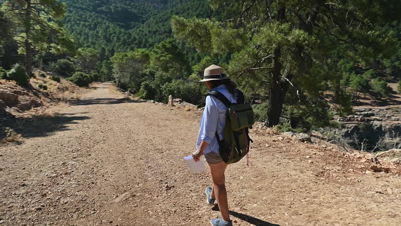 一名女子戴着帽子，背着背包，走在风景秀丽的森林小径上，在树木环绕的大自然中享受着宁静的一天视频素材