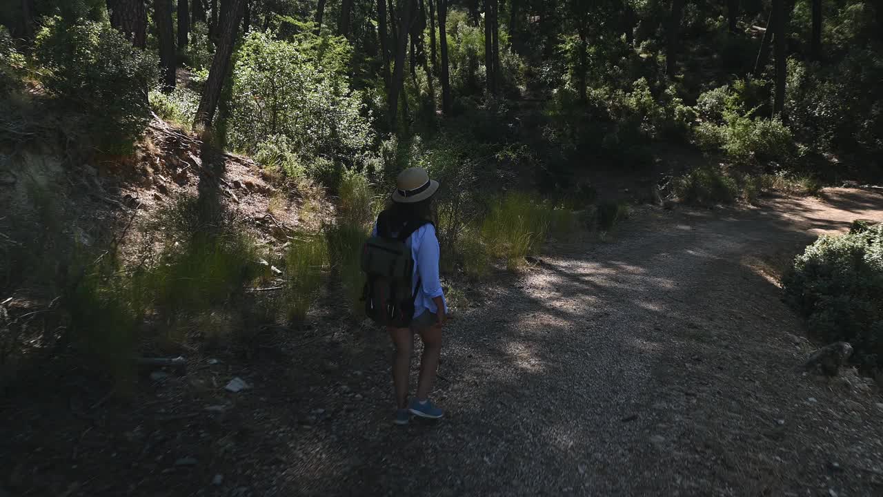 一名妇女戴着帽子背着背包走在森林小路上，享受着大自然包围下的宁静徒步旅行。阳光透过树木，在小径上投下柔和的阴影。视频素材