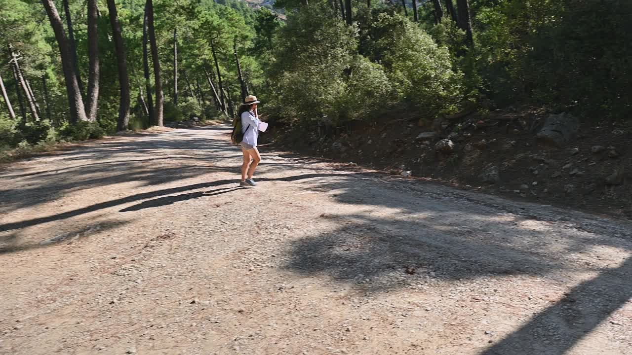 在阳光明媚的日子里，一名女子沿着风景秀丽的森林小径徒步旅行，沐浴在大自然的美景中。独自探索户外的女性冒险家，穿着随意，背着背包，戴着帽子。视频素材