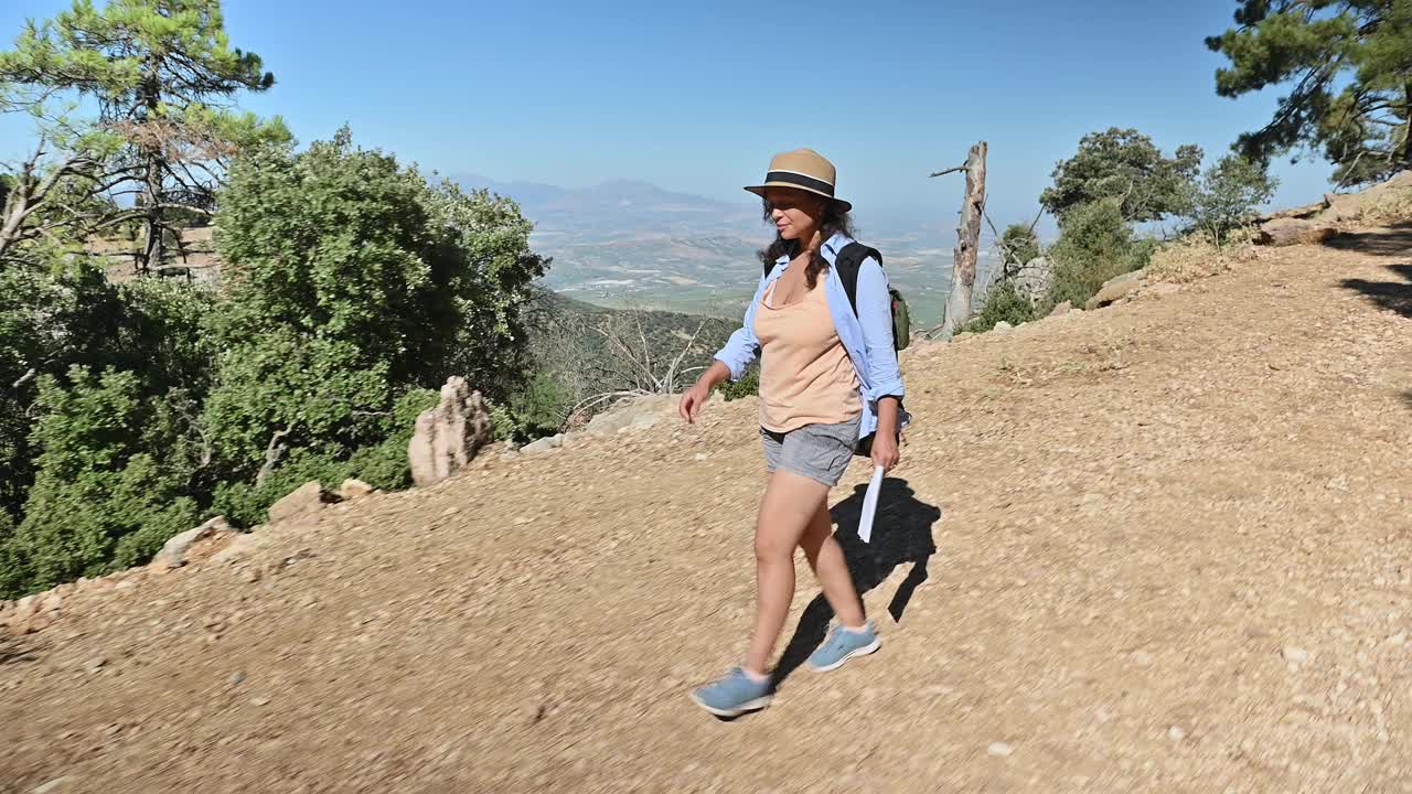 在阳光明媚的日子里，在风景优美的森林小径上徒步旅行的女人，戴着帽子，背着背包，穿着休闲的夏装，享受着大自然，表达着自由和冒险视频素材